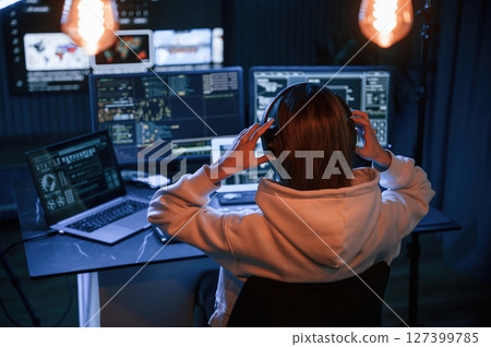Wearing headphones. Young professional female hacker is indoors by computer with lot of information on displays Wearing headphones. Young professional female hacker is indoors by computer with lot of information on displays 127399785