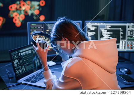 Taking of headphones. Young professional female hacker is indoors by computer with lot of information on displays 127399804