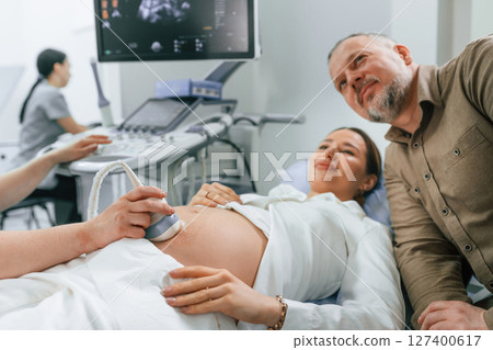 Husband is supporting his wife. Pregnant woman is lying down in the hospital, doctor does ultrasound Husband is supporting his wife. Pregnant woman is lying down in the hospital, doctor does ultrasound 127400617