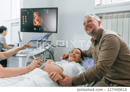 Husband is supporting his wife. Pregnant woman is lying down in the hospital, doctor does ultrasound Husband is supporting his wife. Pregnant woman is lying down in the hospital, doctor does ultrasound 127400618