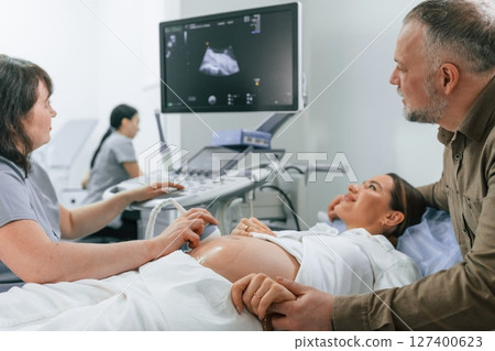 Happy husband is sitting and supporting his wife. Pregnant woman is lying down in the hospital, doctor does ultrasound Happy husband is sitting and supporting his wife. Pregnant woman is lying down in the hospital, doctor does ultrasound 127400623