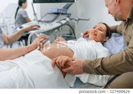 Happy husband is sitting and supporting his wife. Pregnant woman is lying down in the hospital, doctor does ultrasound Happy husband is sitting and supporting his wife. Pregnant woman is lying down in the hospital, doctor does ultrasound 127400626