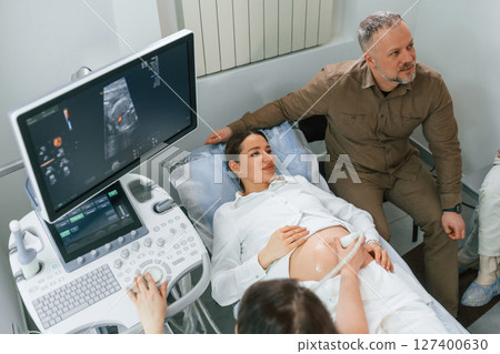 Man is sitting with his wife. Pregnant woman is lying down in the hospital, doctor does ultrasound Man is sitting with his wife. Pregnant woman is lying down in the hospital, doctor does ultrasound 127400630