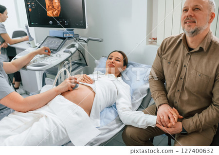 Man is sitting with his wife. Pregnant woman is lying down in the hospital, doctor does ultrasound 127400632