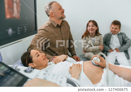 Husband and kids are sitting together. Pregnant woman is lying down in the hospital, doctor does ultrasound 127400675