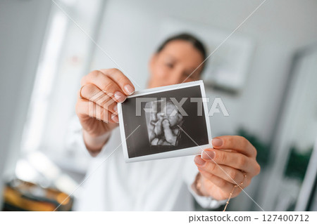 Standing and smiling. Woman in white coat is holding ultrasound picture of baby photo Standing and smiling. Woman in white coat is holding ultrasound picture of baby photo 127400712