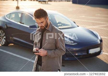 With smartphone. Man is standing near his electric car outdoors 127401692