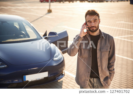 Front view. Man is standing near his electric car outdoors Front view. Man is standing near his electric car outdoors 127401702