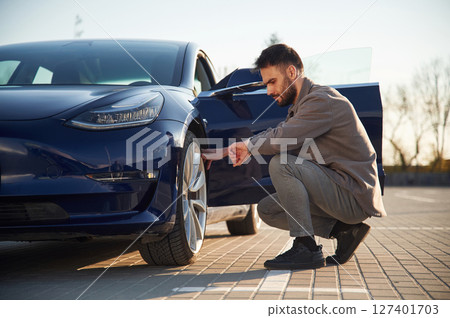 Sitting and checking the wheel. Man is standing near his electric car outdoors Sitting and checking the wheel. Man is standing near his electric car outdoors 127401703