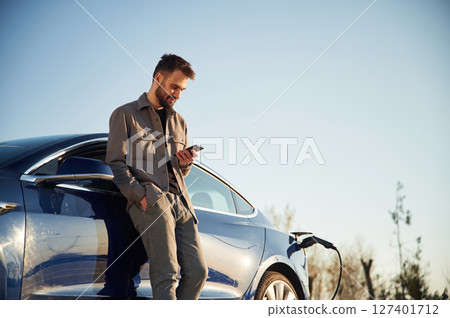 Holding smartphone in hand. Man is standing near his electric car outdoors Holding smartphone in hand. Man is standing near his electric car outdoors 127401712