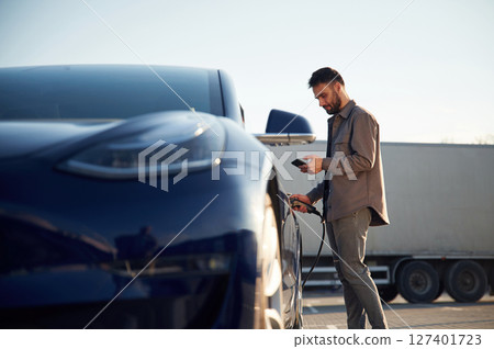 View from the side. Charging the automobile. Man is standing near his electric car outdoors View from the side. Charging the automobile. Man is standing near his electric car outdoors 127401723