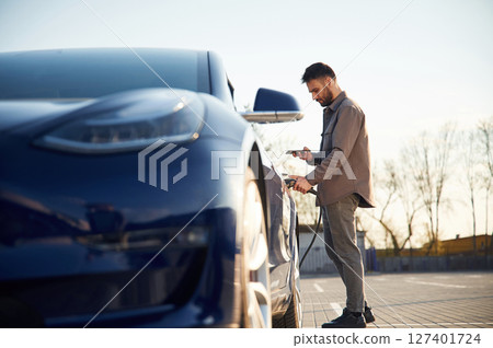 View from the side. Charging the automobile. Man is standing near his electric car outdoors View from the side. Charging the automobile. Man is standing near his electric car outdoors 127401724