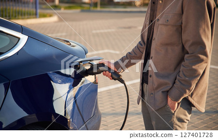 Blue colored automobile. Putting the charger in. Man is standing near his electric car outdoors Blue colored automobile. Putting the charger in. Man is standing near his electric car outdoors 127401728