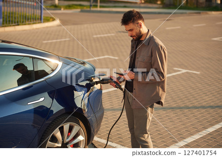 Bored, waiting for automobile to charge. With smartphone. Man is standing near his electric car outdoors Bored, waiting for automobile to charge. With smartphone. Man is standing near his electric car outdoors 127401754
