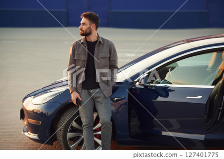 Blue colored building behind. Man is standing near his electric car outdoors Blue colored building behind. Man is standing near his electric car outdoors 127401775