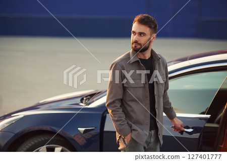 Blue colored building behind. Man is standing near his electric car outdoors Blue colored building behind. Man is standing near his electric car outdoors 127401777