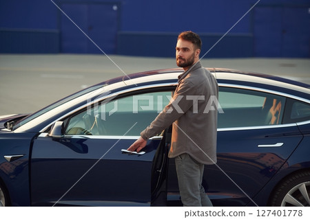 Blue colored building behind. Man is standing near his electric car outdoors Blue colored building behind. Man is standing near his electric car outdoors 127401778