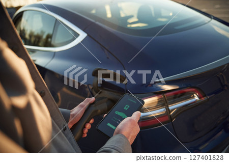 Mobile app with charging data. Man is standing near his electric car outdoors Mobile app with charging data. Man is standing near his electric car outdoors 127401828