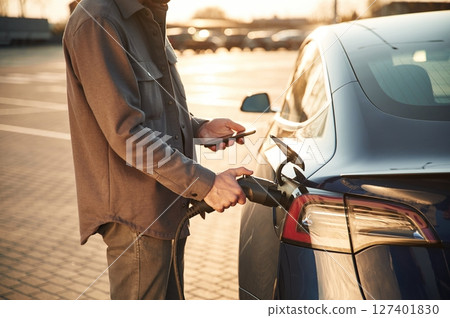 Grey colored formal clothes. Man is standing near his electric car outdoors Grey colored formal clothes. Man is standing near his electric car outdoors 127401830