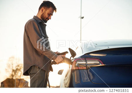 In the beautiful sunlight. Man is standing near his electric car outdoors In the beautiful sunlight. Man is standing near his electric car outdoors 127401831