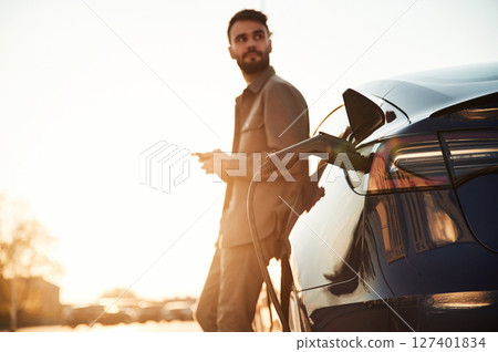 In the beautiful sunlight. Man is standing near his electric car outdoors In the beautiful sunlight. Man is standing near his electric car outdoors 127401834