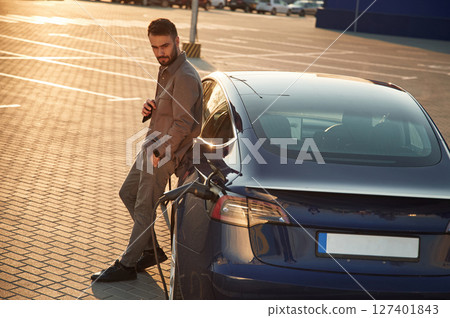 On the charging station. Man is standing near his electric car outdoors On the charging station. Man is standing near his electric car outdoors 127401843