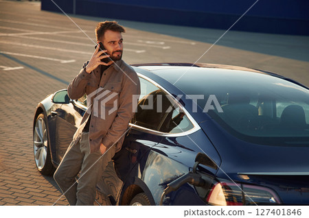Leaning on the automobile and holding smartphone. On the charging station. Man is standing near his electric car 127401846