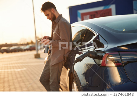 Leaning on the automobile and holding smartphone. On the charging station. Man is standing near his electric car Leaning on the automobile and holding smartphone. On the charging station. Man is standing near his electric car 127401848