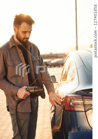 On the parking lot. Charging the automobile. Man is standing near his electric car outdoors On the parking lot. Charging the automobile. Man is standing near his electric car outdoors 127401858