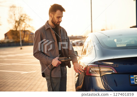 On the parking lot. Charging the automobile. Man is standing near his electric car outdoors 127401859