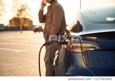 Talking by phone. Man is standing near his electric car outdoors Talking by phone. Man is standing near his electric car outdoors 127401861