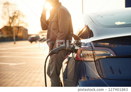 Talking by phone. Man is standing near his electric car outdoors Talking by phone. Man is standing near his electric car outdoors 127401863