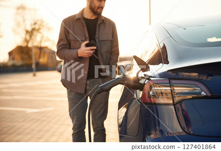 On the parking lot. Charging the automobile. Man is standing near his electric car outdoors On the parking lot. Charging the automobile. Man is standing near his electric car outdoors 127401864