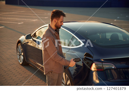 Putting charger into socket. Man is standing near his electric car outdoors Putting charger into socket. Man is standing near his electric car outdoors 127401879