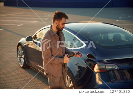 Putting charger into socket. Man is standing near his electric car outdoors Putting charger into socket. Man is standing near his electric car outdoors 127401880