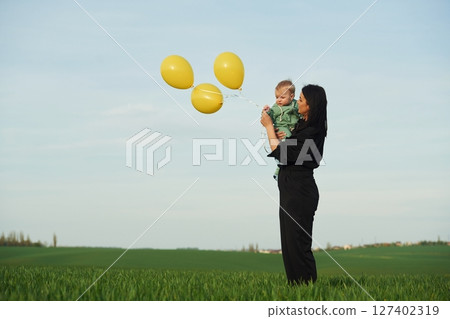 With yellow colored balloons. Mother with her little baby son is outdoors on the agricultural field 127402319