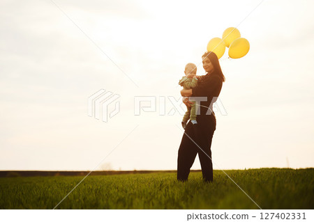 Holding yellow colored balloons in hands. Mother with her little baby son is outdoors on the agricultural field 127402331