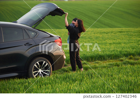 Standing near car, closing trunk. Mother with her little baby son is outdoors on the agricultural field 127402346
