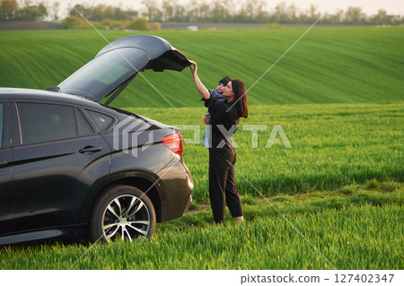 Standing near car, closing trunk. Mother with her little baby son is outdoors on the agricultural field Standing near car, closing trunk. Mother with her little baby son is outdoors on the agricultural field 127402347