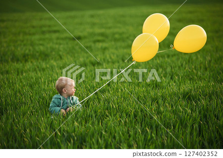 With yellow balloons in hands. Cute little baby boy is in the agricultural field at daytime 127402352