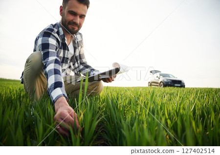 Car is behind. With notepad and wheat in hands. Handsome young man is on agricultural field 127402514