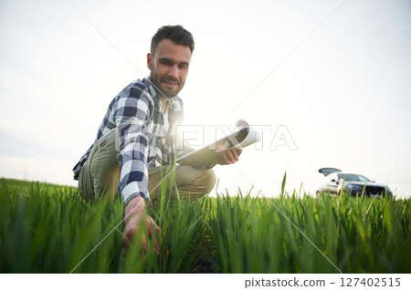 Car is behind. With notepad and wheat in hands. Handsome young man is on agricultural field 127402515