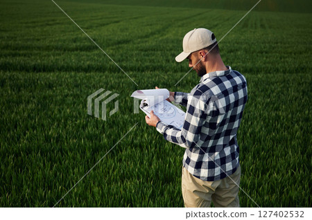 Standing, holding document. In cap. Handsome young man is on agricultural field Standing, holding document. In cap. Handsome young man is on agricultural field 127402532