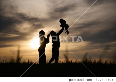 Holding little baby in hands. Silhouettes of father and mother that are outdoors against sunset dramatic sky in the field Holding little baby in hands. Silhouettes of father and mother that are outdoors against sunset dramatic sky in the field 127402593
