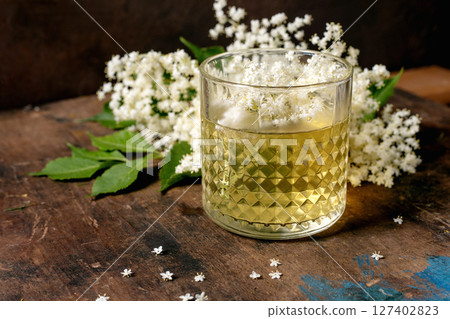 Crystal glass with elderflower cocktail decorated with white elderflower blossoms on rustic wooden table. Natural herbal drink with floral garnish. 127402823