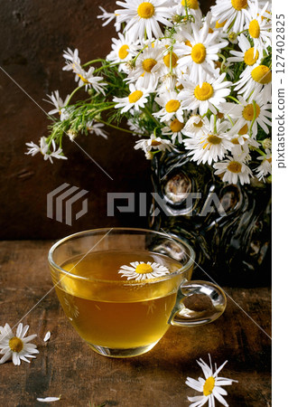 Glass cup herbal chamomile tea with white daisy flowers in vintage vase on wooden table. Natural wellness drink concept with fresh blooms decoration. Glass cup herbal chamomile tea with white daisy flowers in vintage vase on wooden table. Natural wellness drink concept with fresh blooms decoration. 127402825