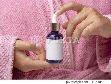 Woman in pink bathrobe with blue dropper bottle with label in hands closeup, cosmetic mockup 127404100