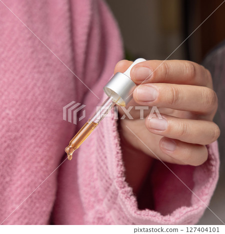 Young woman in pink bathrobe dropping essential oil or serum from a pipette closeup Young woman in pink bathrobe dropping essential oil or serum from a pipette closeup 127404101