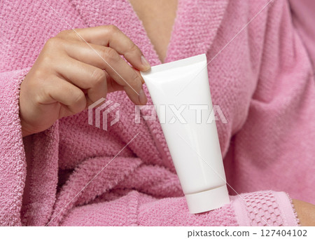 Closeup of young woman in pink bathrobe holding squeeze cream tube in hands, cosmetic mockup 127404102