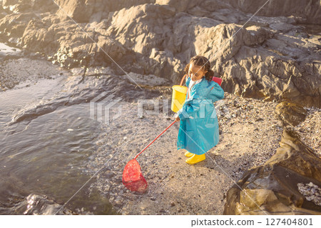 Girl, beach and fishing with net in rock pool, water or ocean with boots, bucket and coat in sunshine. Black child, sea and outdoor for fish, marine life or animal in nature, learning and adventure 127404801